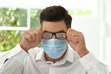 Man wiping foggy glasses caused by wearing medical mask indoors, closeup