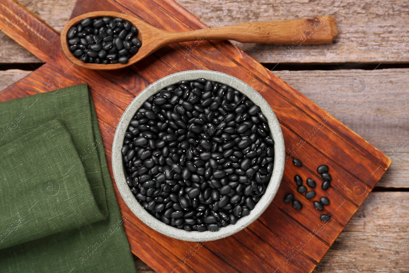 Photo of Bowl and spoon of raw black beans on wooden table, flat lay