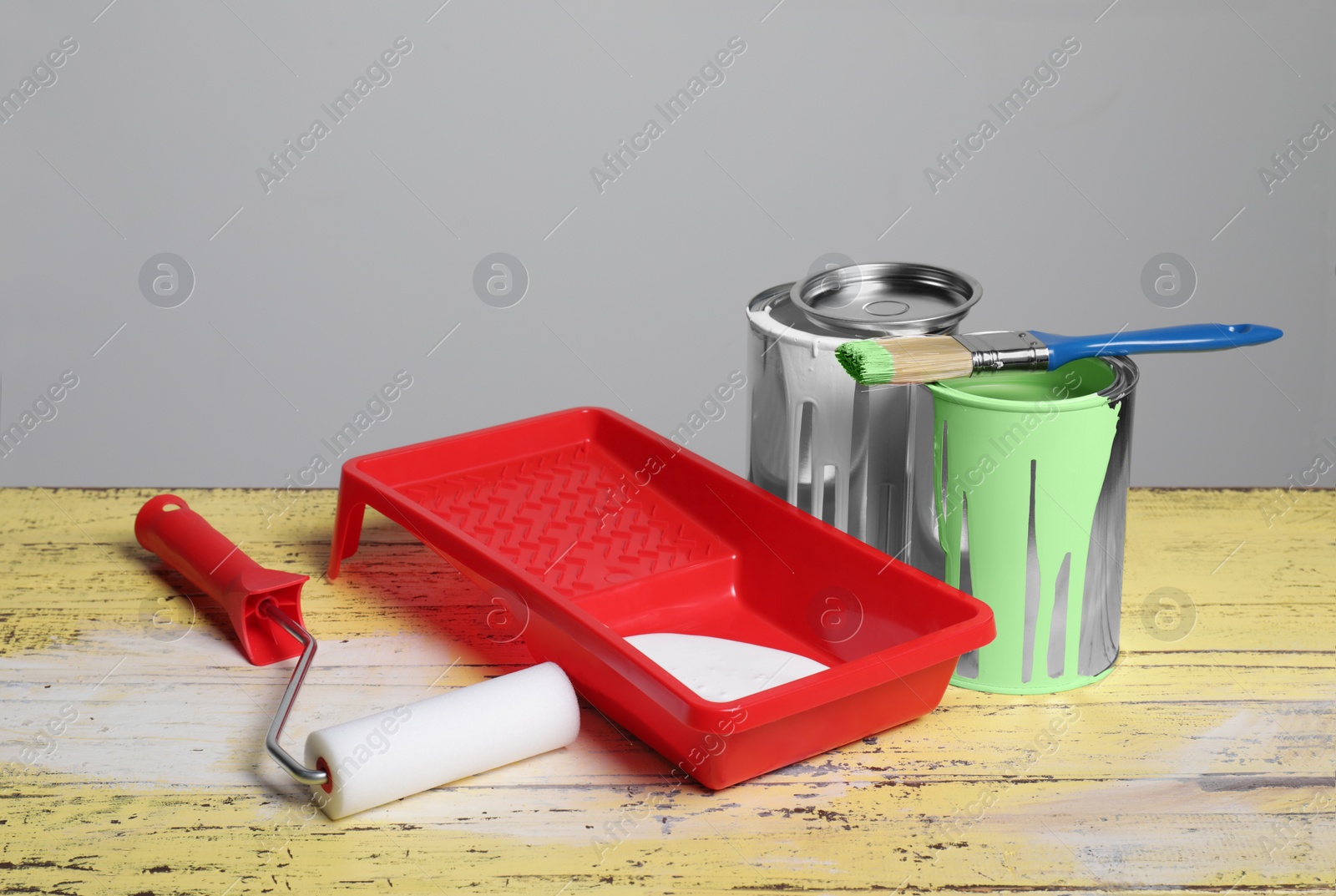 Photo of Cans of paints, brush, roller and tray on yellow wooden table