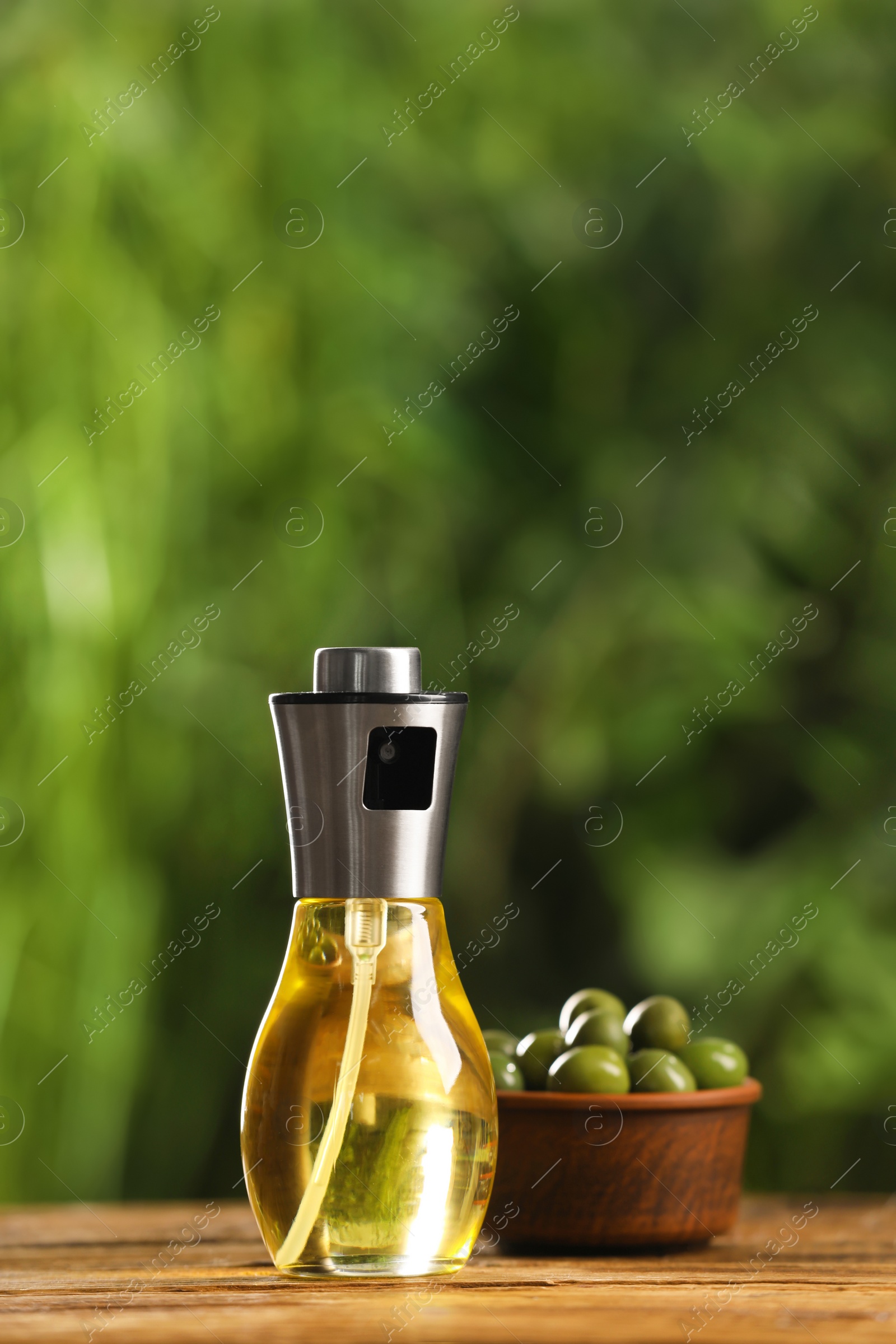 Photo of Spray bottle with cooking oil and olives on wooden table against blurred background