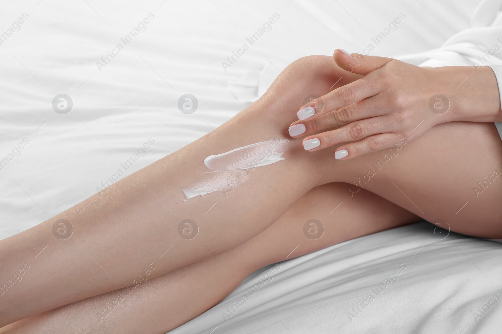 Photo of Woman applying body cream onto her smooth legs on bed, closeup