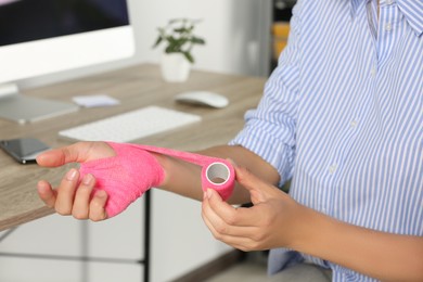 Photo of Young woman applying medical bandage onto hand at workplace, closeup