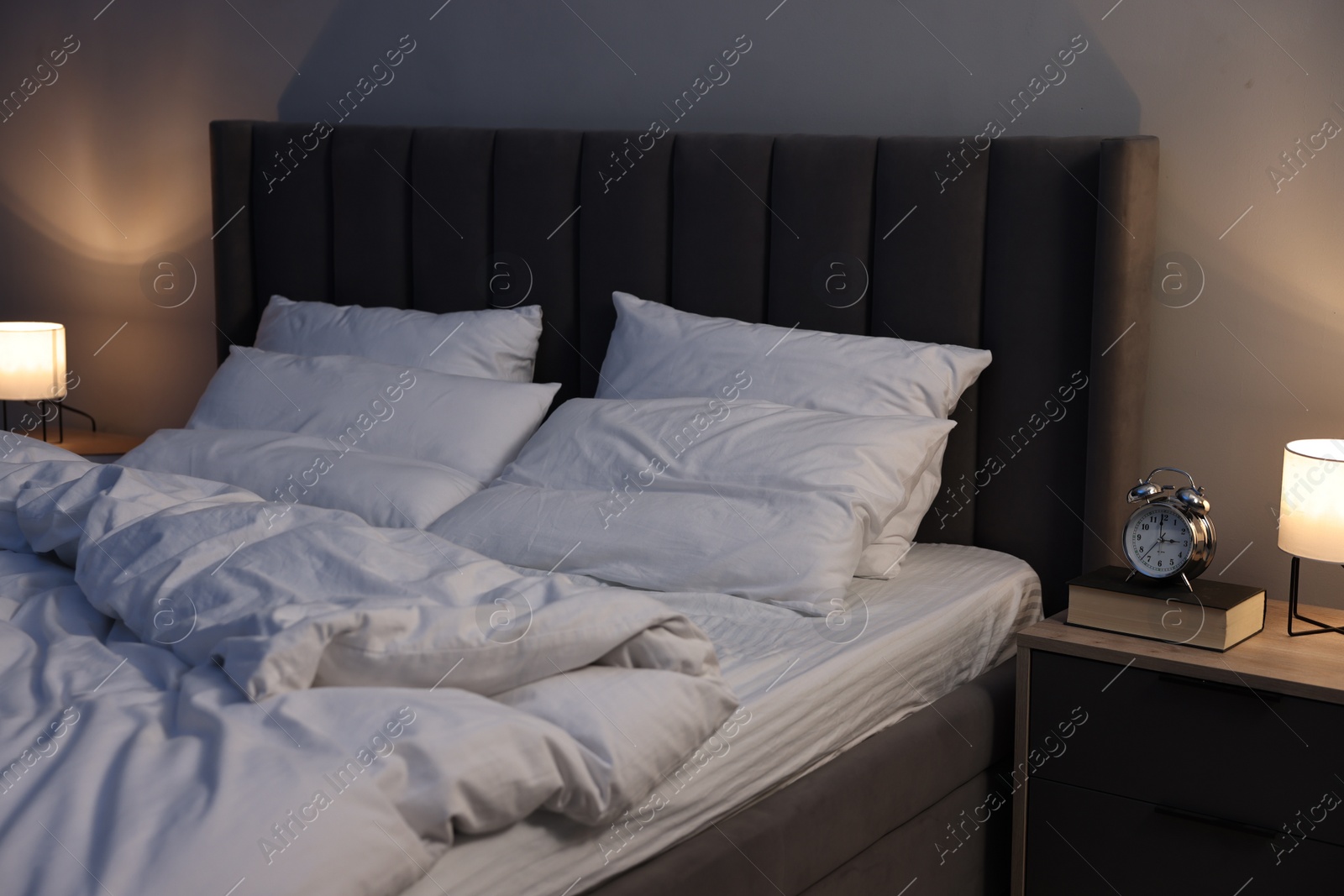 Photo of Nightlight, alarm clock and book on bedside table near bed indoors