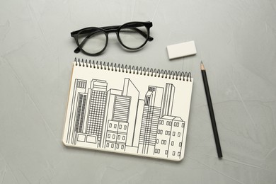 Sketch of buildings in notebook, pencil, eraser and glasses on grey table, flat lay