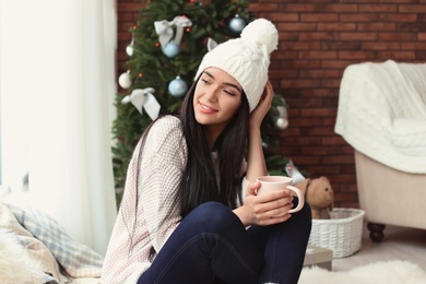 Beautiful young woman in hat with cup of drink at home. Christmas celebration