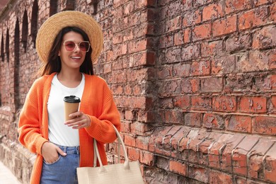 Photo of Young woman with stylish bag and cup of hot drink near red brick wall outdoors, space for text