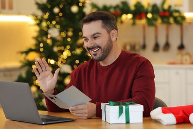 Celebrating Christmas online with exchanged by mail presents. Happy man with greeting card waving hello during video call on laptop at home