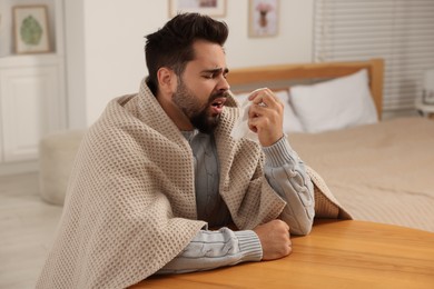 Photo of Sick man wrapped in blanket with tissue at wooden table indoors. Cold symptoms