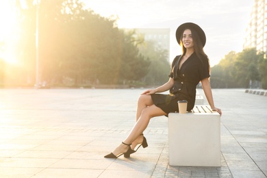 Beautiful young woman in stylish black dress and hat with cup of coffee on city street