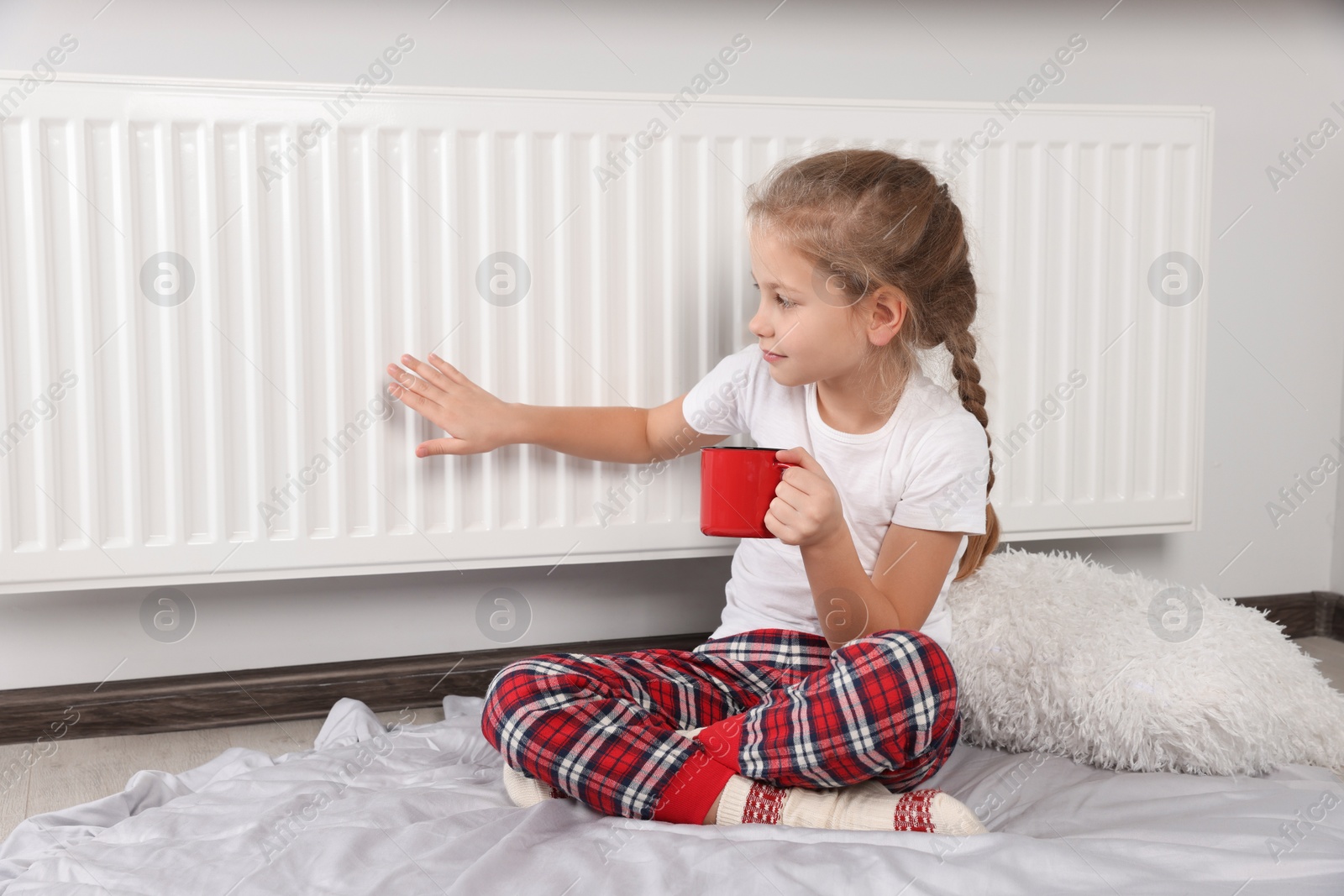 Photo of Little girl with cup of hot drink near heating radiator indoors