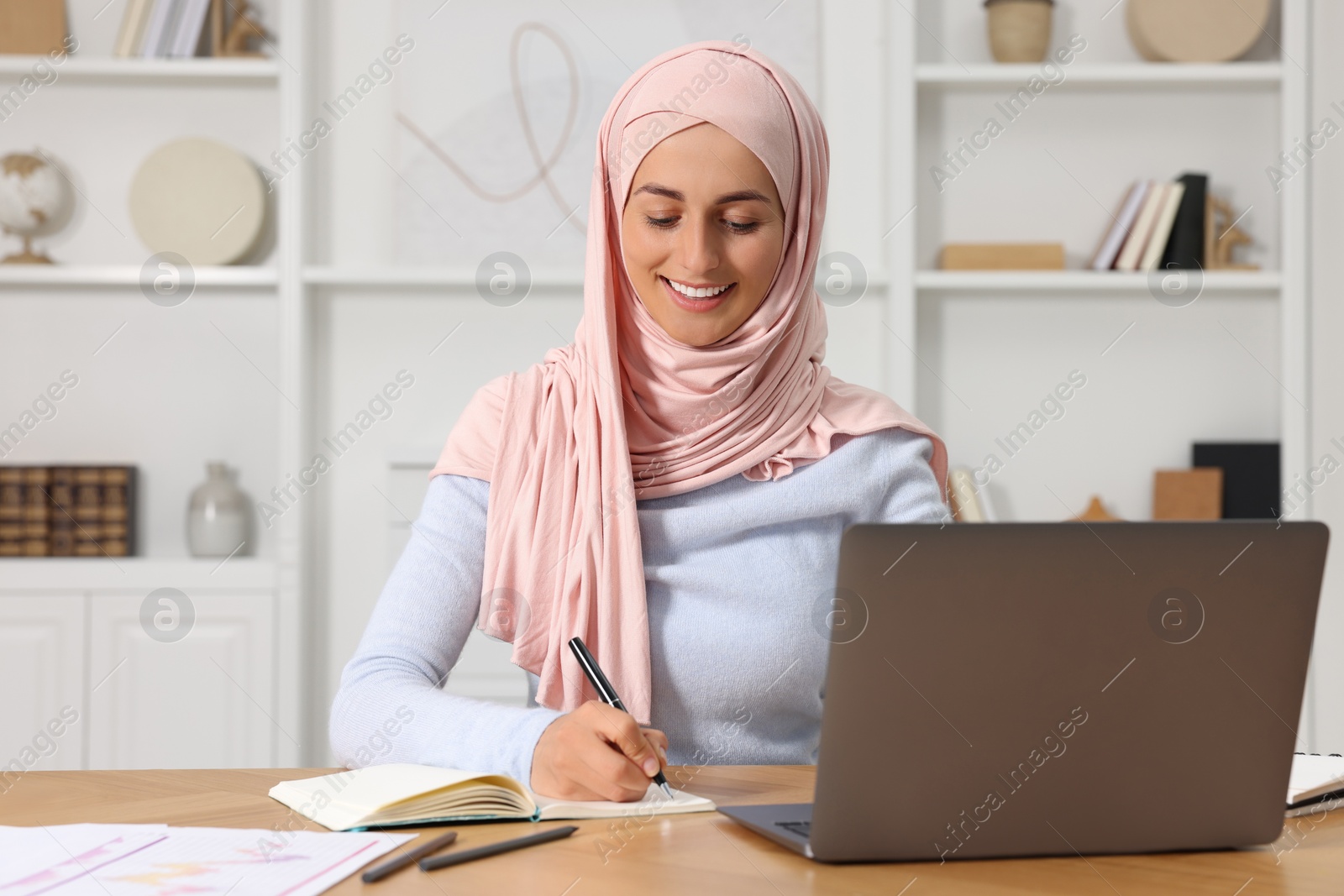 Photo of Muslim woman writing notes near laptop at wooden table in room