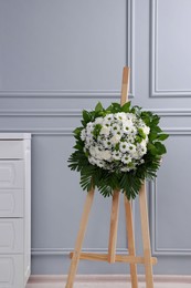 Photo of Funeral wreath of flowers on wooden stand near light grey wall indoors
