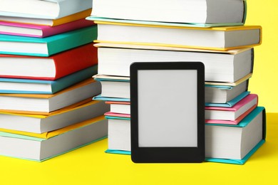 Modern e-book reader and stacks of hard cover books on yellow background, closeup