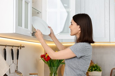 Photo of Beautiful young woman taking plate from kitchen cabinet at home