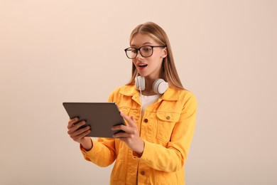 Emotional teenage student with tablet and headphones on beige background