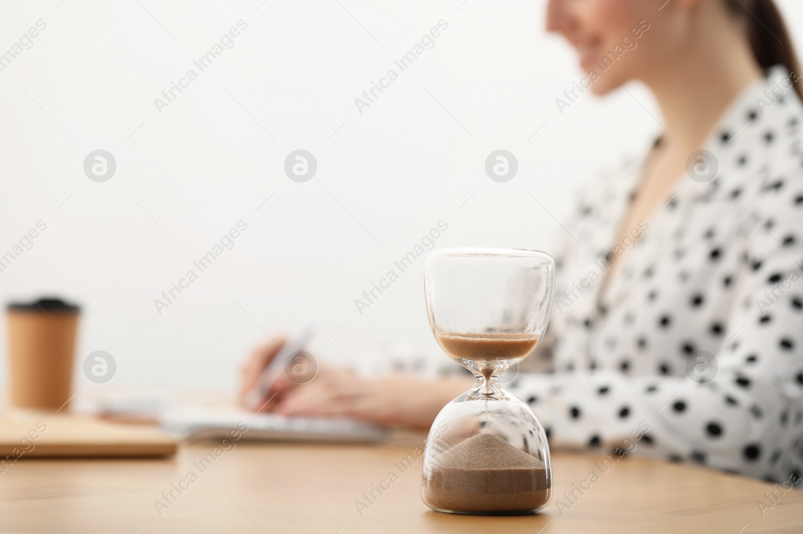 Photo of Hourglass with flowing sand on desk. Woman taking notes while using calculator indoors, selective focus