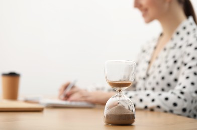 Photo of Hourglass with flowing sand on desk. Woman taking notes while using calculator indoors, selective focus