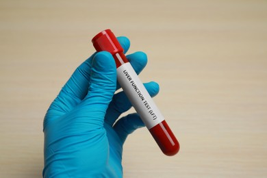 Laboratory worker holding tube with blood sample and label Liver Function Test at beige table, closeup
