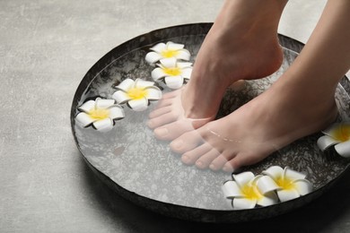 Photo of Woman soaking her feet in bowl with water and flowers on light grey floor, closeup. Spa treatment