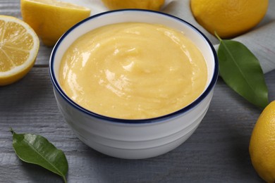 Delicious lemon curd in bowl, fresh citrus fruits and green leaves on grey wooden table, closeup