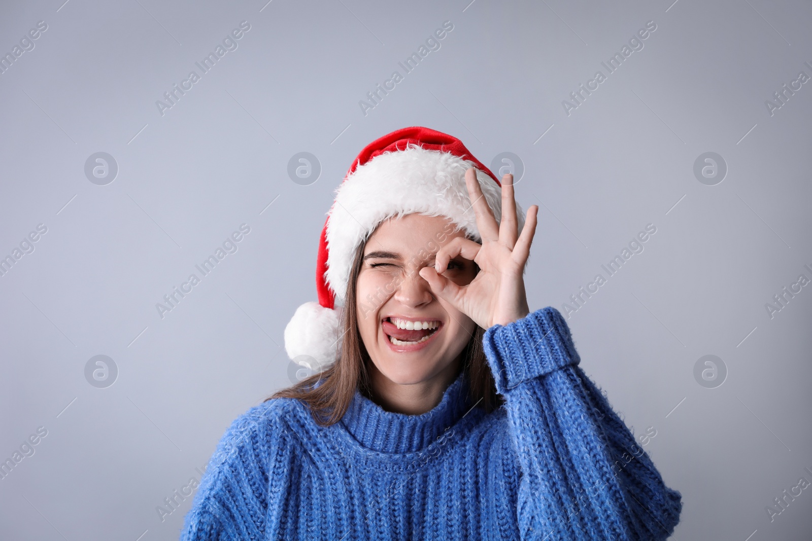 Photo of Playful woman in Santa hat and blue sweater showing OK gesture on grey background