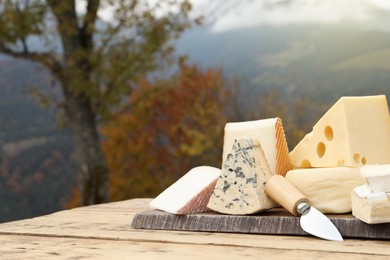 Photo of Different types of delicious cheeses on wooden table against mountain landscape. Space for text