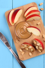 Slices of fresh apple with peanut butter, nuts and knife on light blue wooden table, flat lay