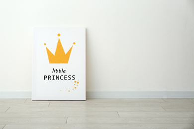 Photo of Adorable picture of crown with words LITTLE PRINCESS on floor near white wall, space for text. Children's room interior element
