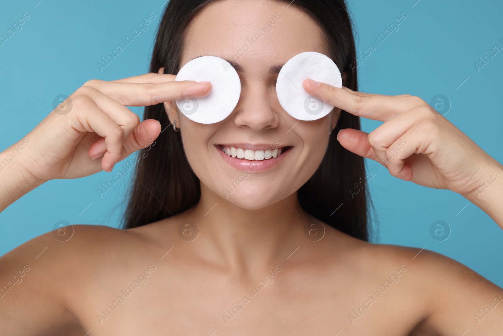 Photo of Young woman covering her eyes with cotton pads on light blue background, closeup