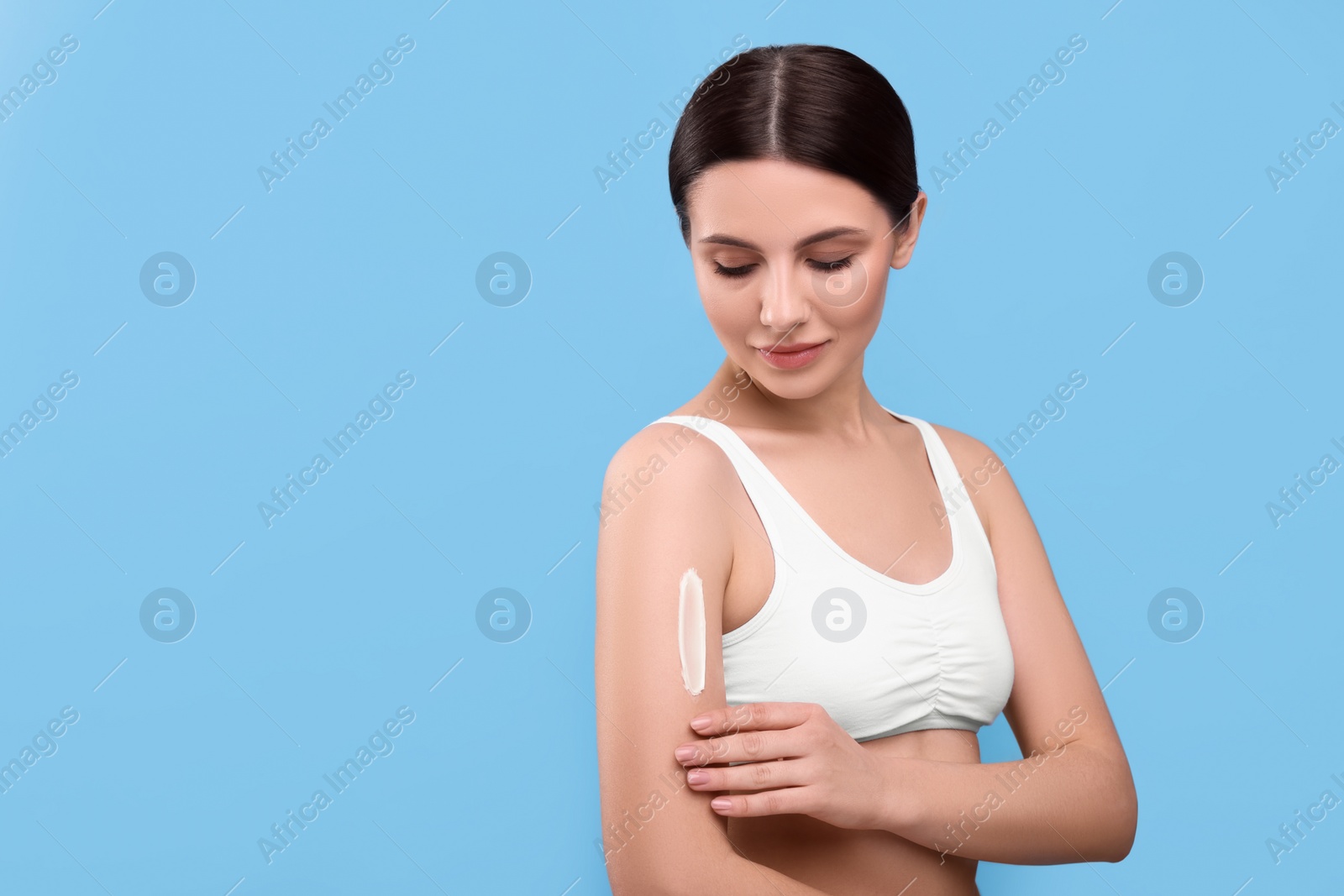 Photo of Beautiful woman with smear of body cream on her arm against light blue background, space for text