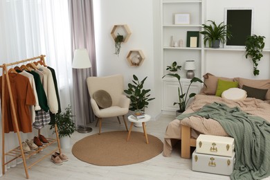 Stylish bedroom with comfortable bed, clothes rack and different houseplants. Interior design