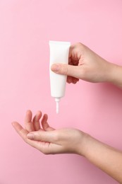 Woman applying cosmetic cream from tube onto her hand on pink background, closeup