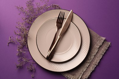 Stylish table setting. Plates, cutlery, napkin and floral decor on purple background, top view