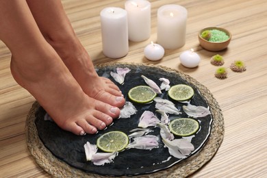 Woman soaking her feet in plate with water, flower petals and lime slices on wooden floor, closeup. Pedicure procedure