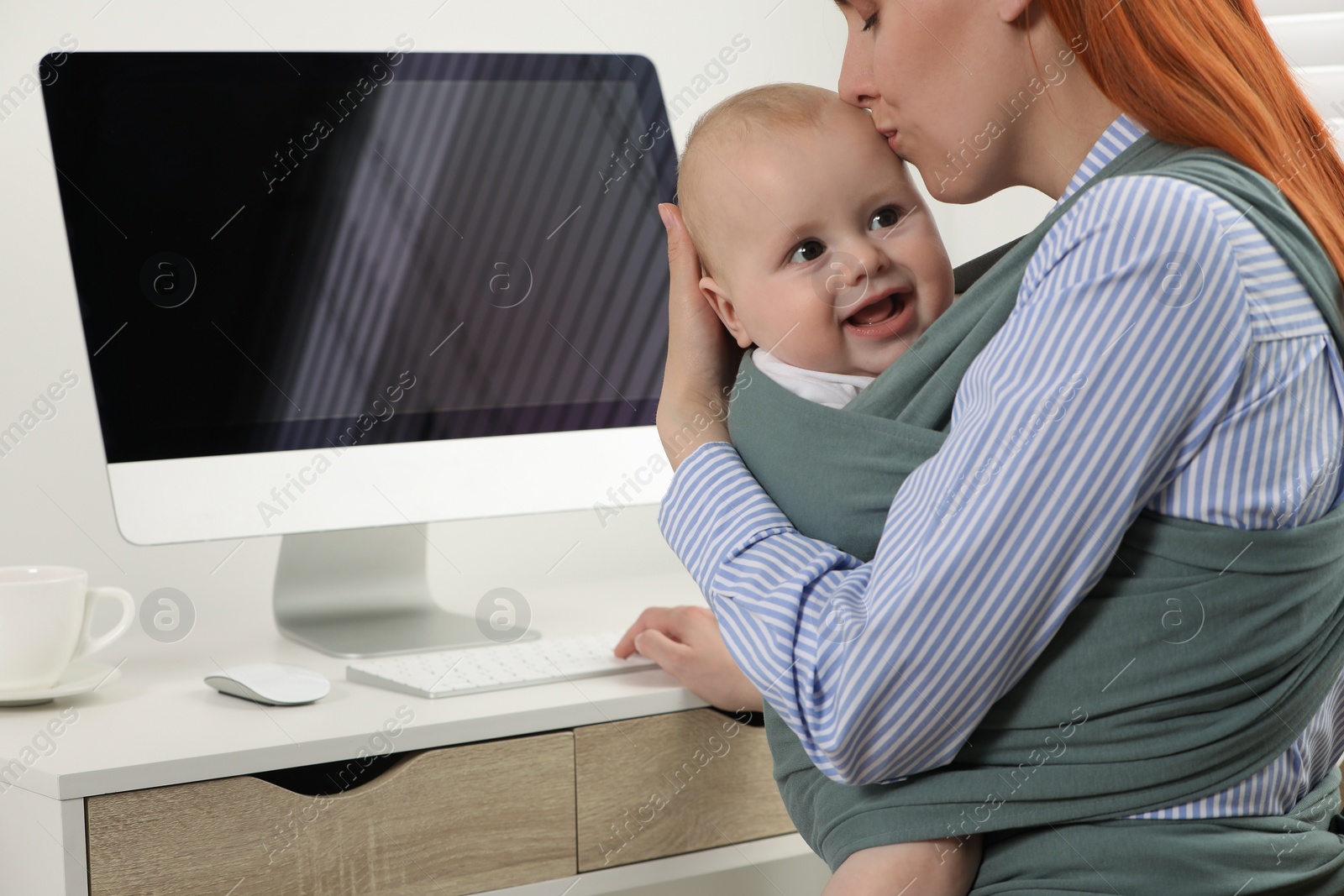 Photo of Mother holding her child in sling (baby carrier) while using computer at workplace