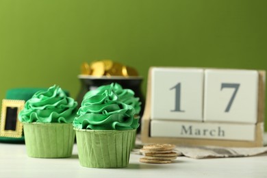 St. Patrick's day party. Tasty cupcakes with green cream, pot of gold, wooden block calendar and leprechaun hat on white table, closeup