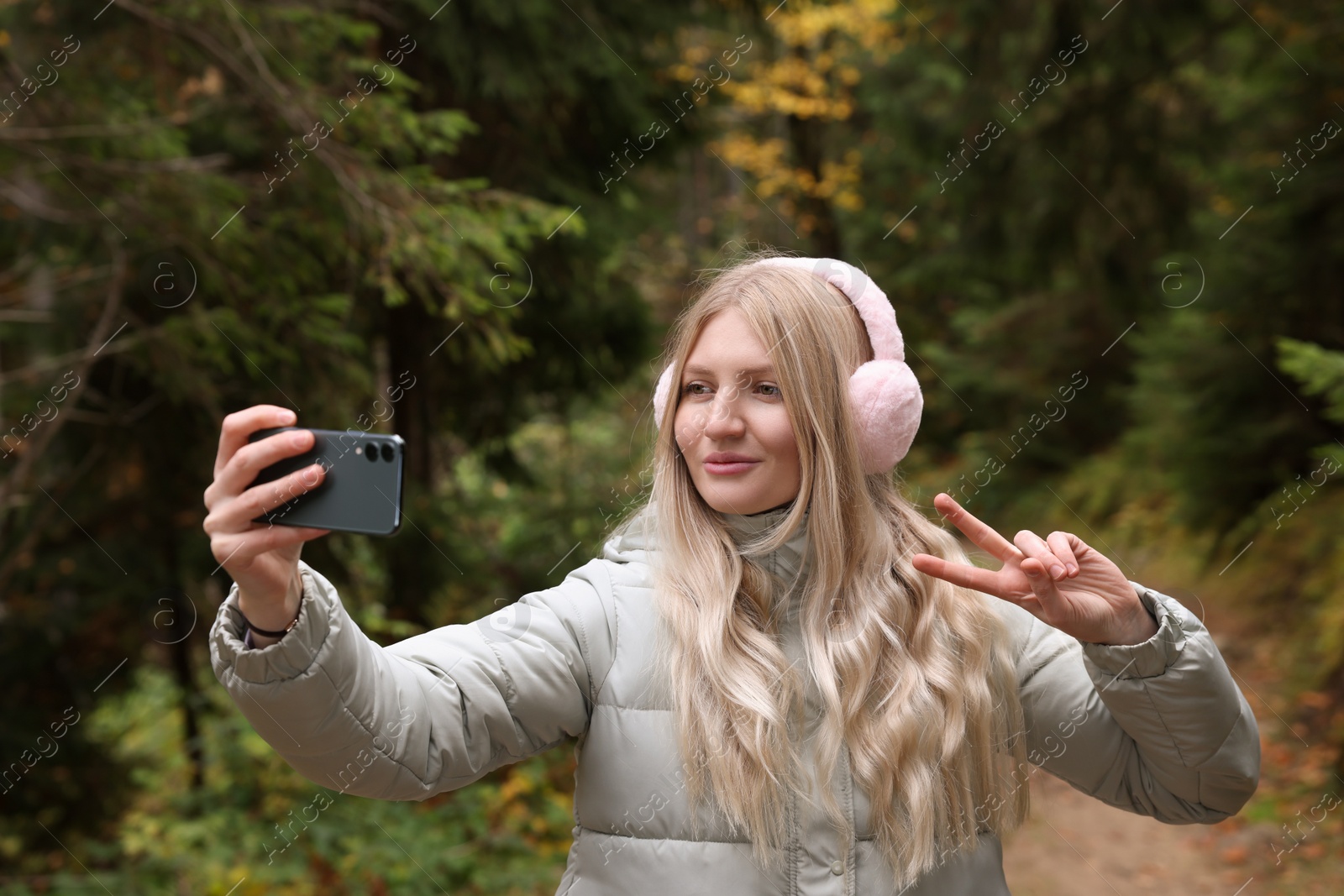 Photo of Young woman in warm earmuffs taking selfie near forest on autumn day
