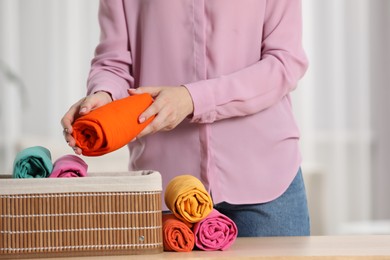 Photo of Woman putting rolled shirt into basket at table indoors, closeup. Organizing clothes