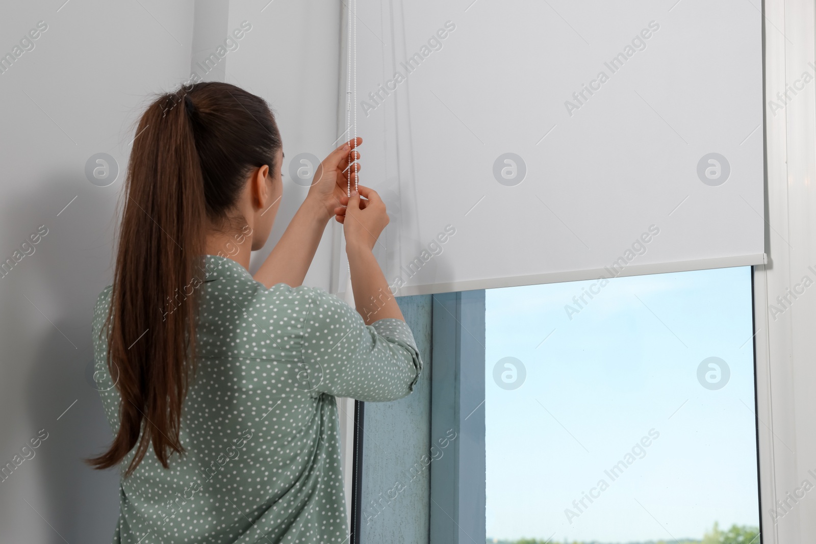 Photo of Woman opening white roller blind on window indoors, back view