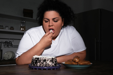 Photo of Depressed overweight woman eating cake in kitchen at night