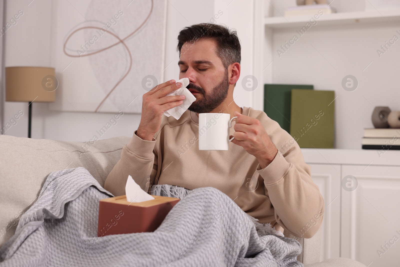 Photo of Sick man with cup of drink and tissue blowing nose on sofa at home. Cold symptoms