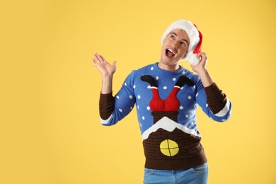 Photo of Portrait of young man in Christmas sweater and Santa hat on yellow background