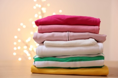 Photo of Stack of folded clothes on wooden table against beige background with blurred lights