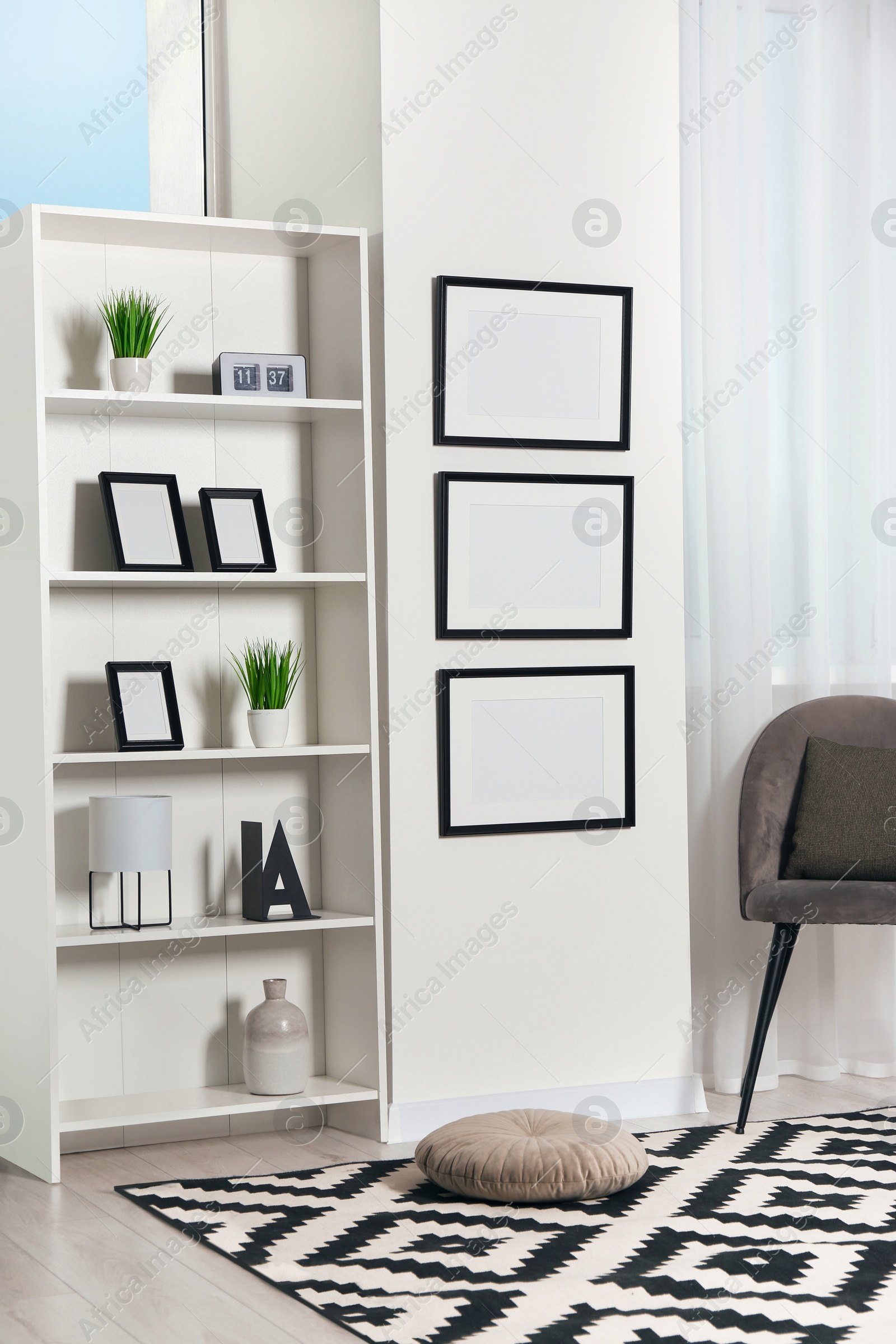 Photo of Stylish room interior with empty frames hanging on white wall near shelving unit