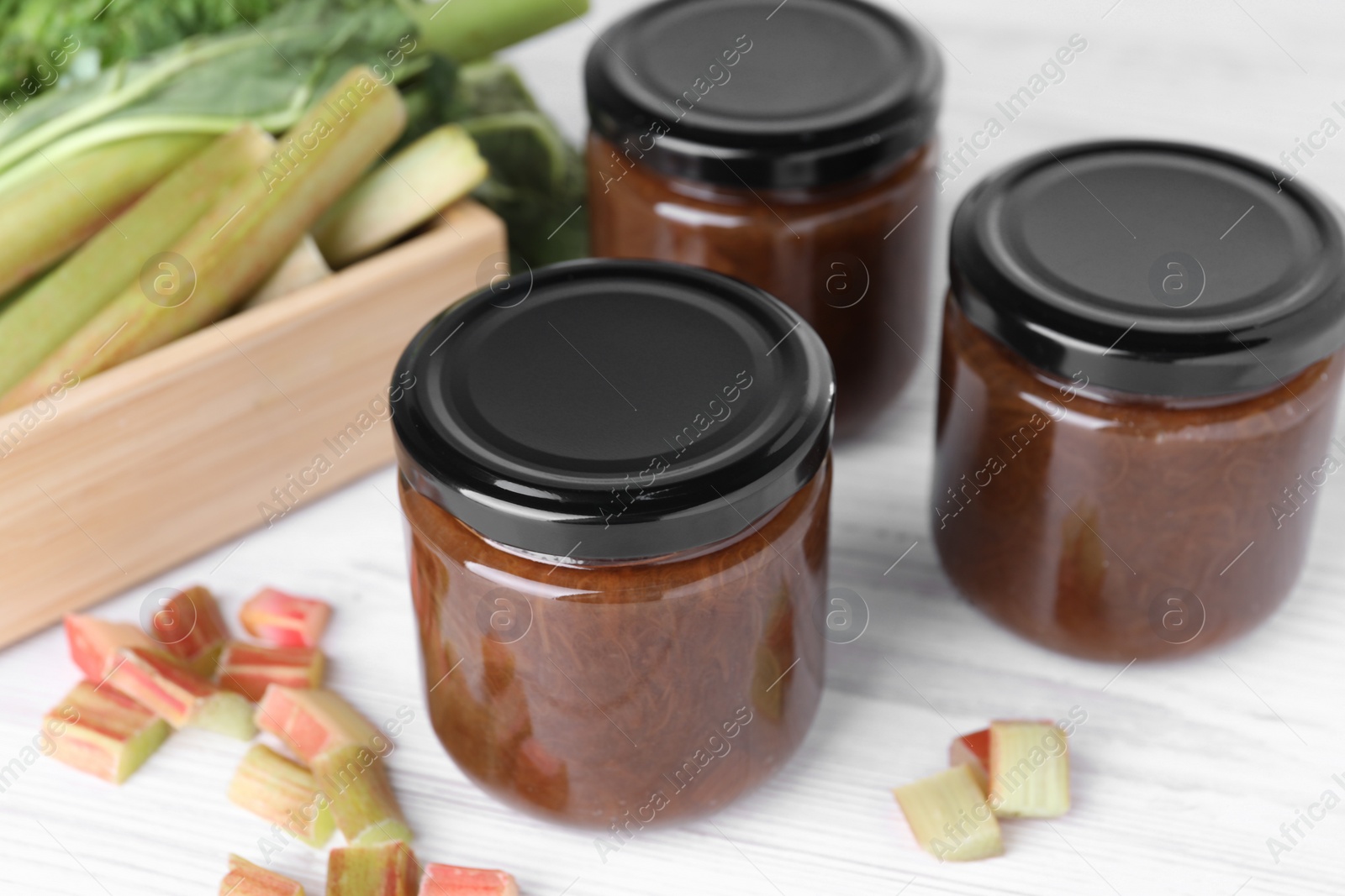 Photo of Jars of tasty rhubarb jam and cut stalks on white wooden table, closeup