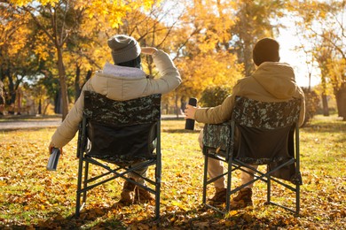 Couple with thermoses sitting in camping chairs outdoors on autumn sunny day, back view