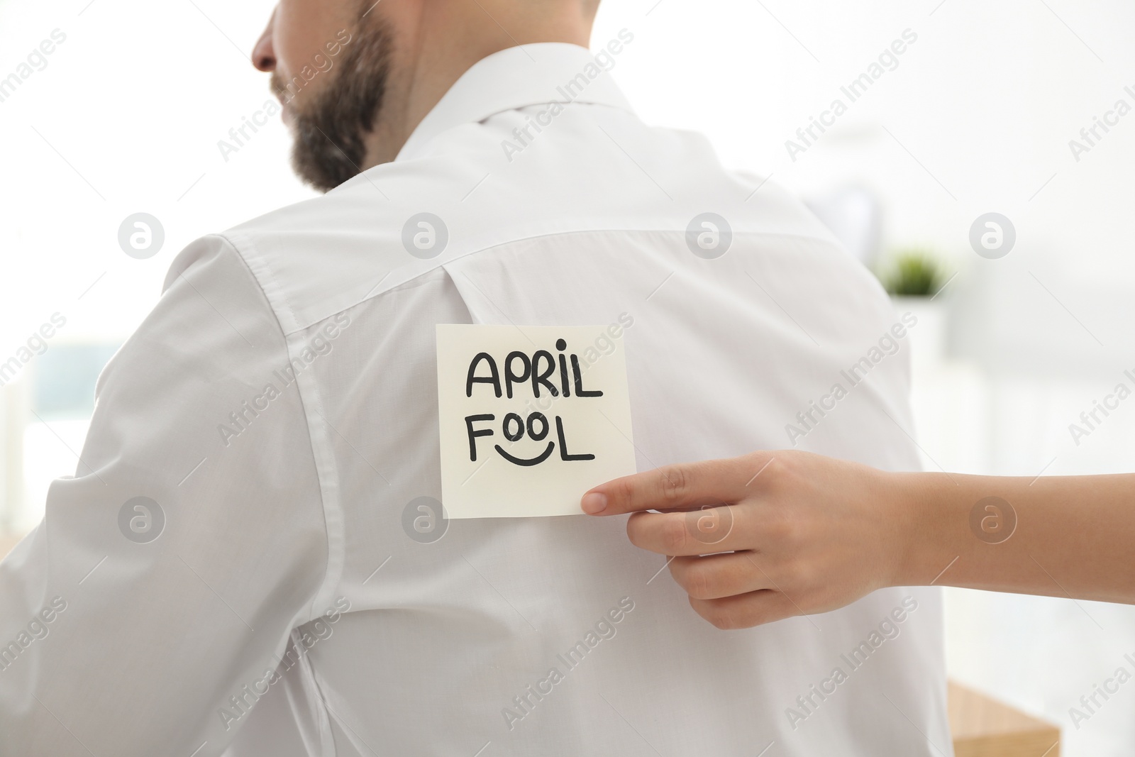 Photo of Woman sticking APRIL FOOL note to colleague's back in office, closeup