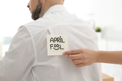 Photo of Woman sticking APRIL FOOL note to colleague's back in office, closeup