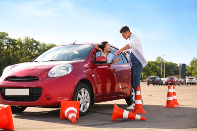 Photo of Stressed young woman in car near instructor and fallen traffic cones outdoors. Failed driving school exam