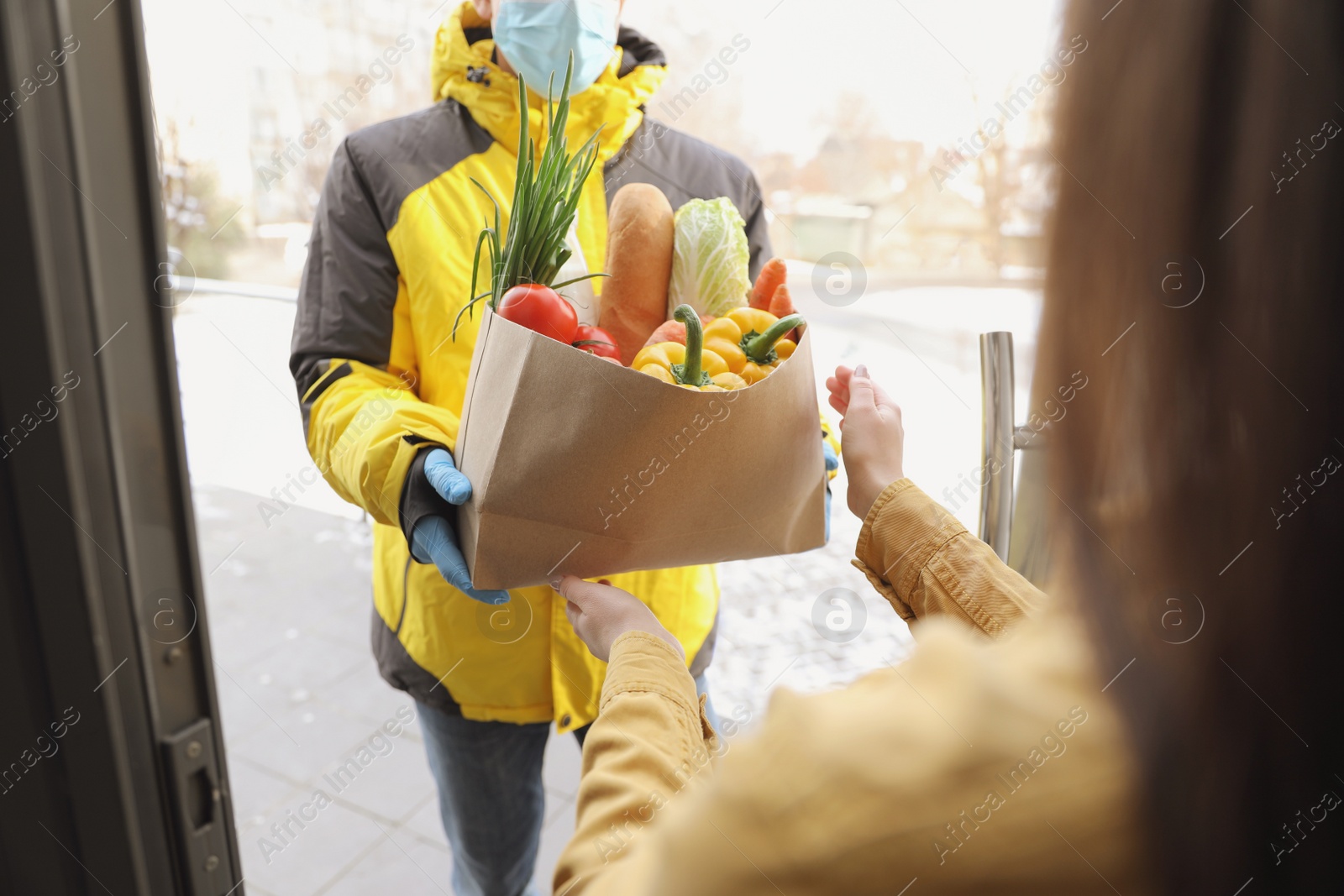 Photo of Courier in medical mask giving paper bag with groceries to woman at doorway, closeup. Delivery service during Covid-19 quarantine